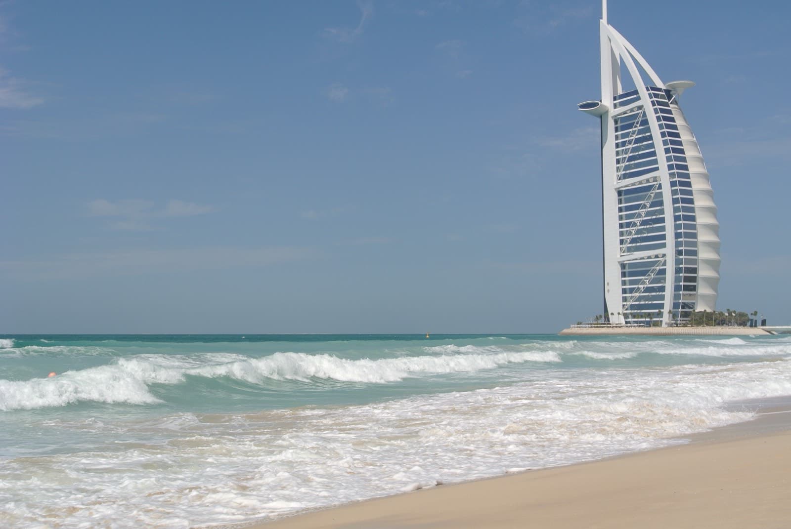 Veřejná pláž v Dubaji s jemným pískem, vlnami a hotelem Burj Al Arab v pozadí